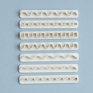 Può includere: Set di otto strumenti per la decorazione di torte in plastica bianca con diversi disegni di bordo. Gli strumenti sono disposti verticalmente su uno sfondo blu chiaro.