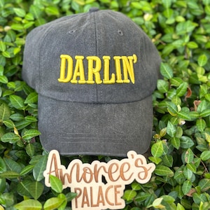 以下が含まれることがあります： 黄色い刺繍で「DARLIN'」の文字が入った濃いグレーの野球帽。 緑の葉を背景に、木製の看板には「Amoree's PALACE」と書かれています。