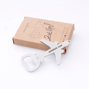 May include: A silver airplane-shaped bottle opener with the text "AIRBUS 320" engraved on the wing. The bottle opener is on a brown cardboard box with the text "Zek Yapp!" and a website address.