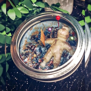 May include: A candle in a silver-rimmed jar with a light blue wax base. The candle features a small figurine, decorative stones, and glitter. A red-topped pin is inserted into the wax. Green leaves are visible in the background.