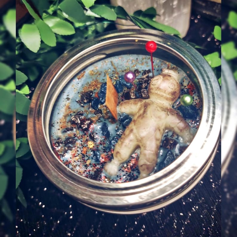 May include: A candle in a silver-rimmed jar with a light blue wax base. The candle features a small figurine, decorative stones, and glitter. A red-topped pin is inserted into the wax. Green leaves are visible in the background.