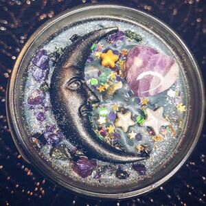 May include: A black crescent moon with a face, a purple heart-shaped crystal, and gold and white stars embedded in a clear candle with a glittery, silver-grey base.