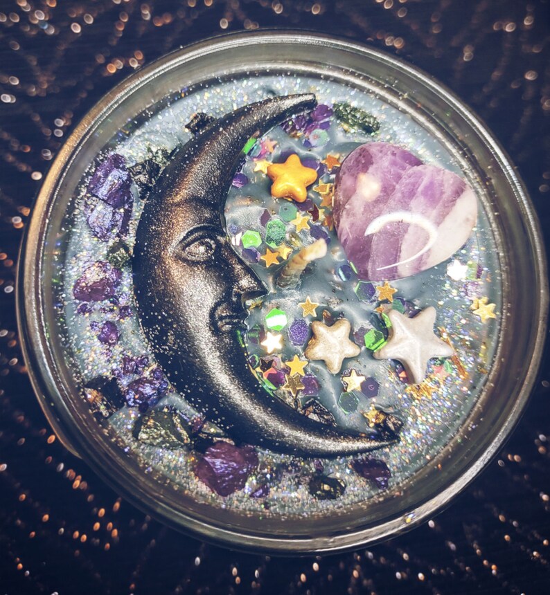 May include: A black crescent moon with a face, a purple heart-shaped crystal, and gold and white stars embedded in a clear candle with a glittery, silver-grey base.