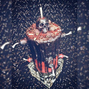 May include: A black candle with a skull on top, surrounded by dried berries and herbs. The candle has a red base and is decorated with bone-shaped accents. It sits on a decorative silver base, creating a gothic or Halloween theme.