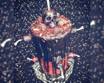 May include: A black candle with a skull on top, surrounded by dried berries and herbs. The candle has a red base and is decorated with bone-shaped accents. It sits on a decorative silver base, creating a gothic or Halloween theme.