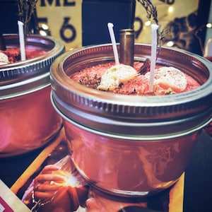 May include: Two glass jars with red wax and white decorative elements. The jars have silver lids and are topped with white wicks. The jars are sitting on a surface with a record player and a book.