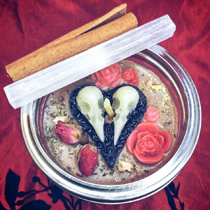 Puede incluir: Un tarro de metal plateado con una tapa de vidrio transparente que contiene una vela rosa y blanca con un dise&ntilde;o de calavera. La vela est&aacute; rodeada de flores y hierbas secas.