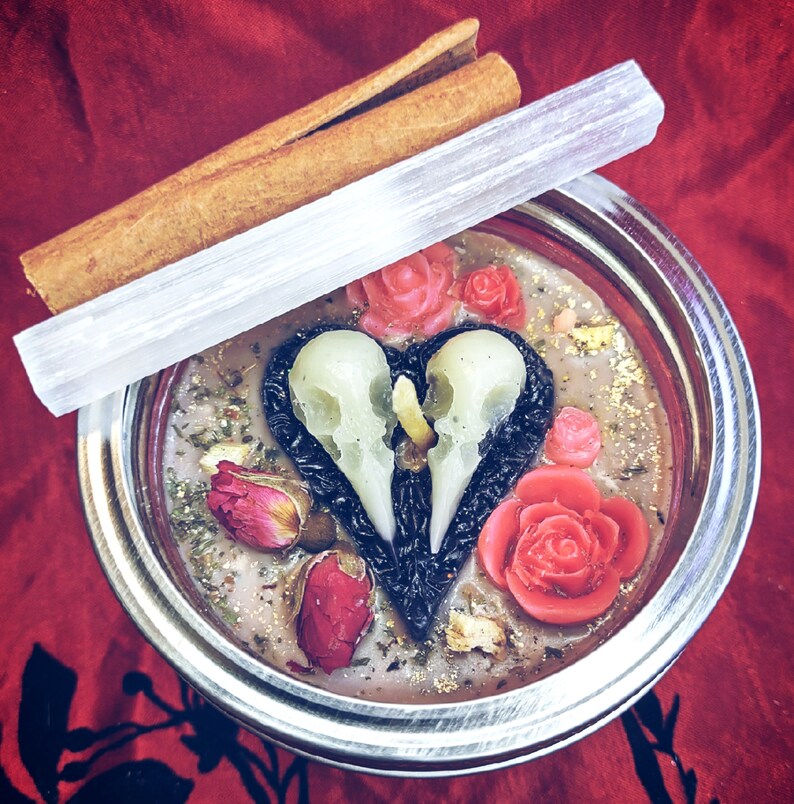 Puede incluir: Un tarro de metal plateado con una tapa de vidrio transparente que contiene una vela rosa y blanca con un dise&ntilde;o de calavera. La vela est&aacute; rodeada de flores y hierbas secas.
