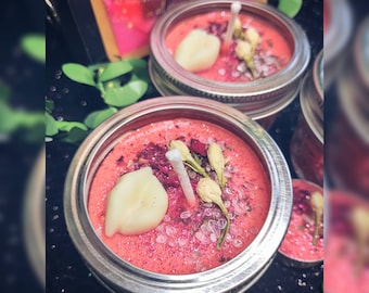 May include: Three pink jar candles with white wicks and white wax accents. The candles are topped with dried flowers and glitter. The jars have silver lids.