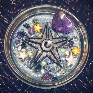 May include: A teal blue candle with a five-pointed star, purple amethyst crystal, and gold glitter. The candle is in a clear glass jar.