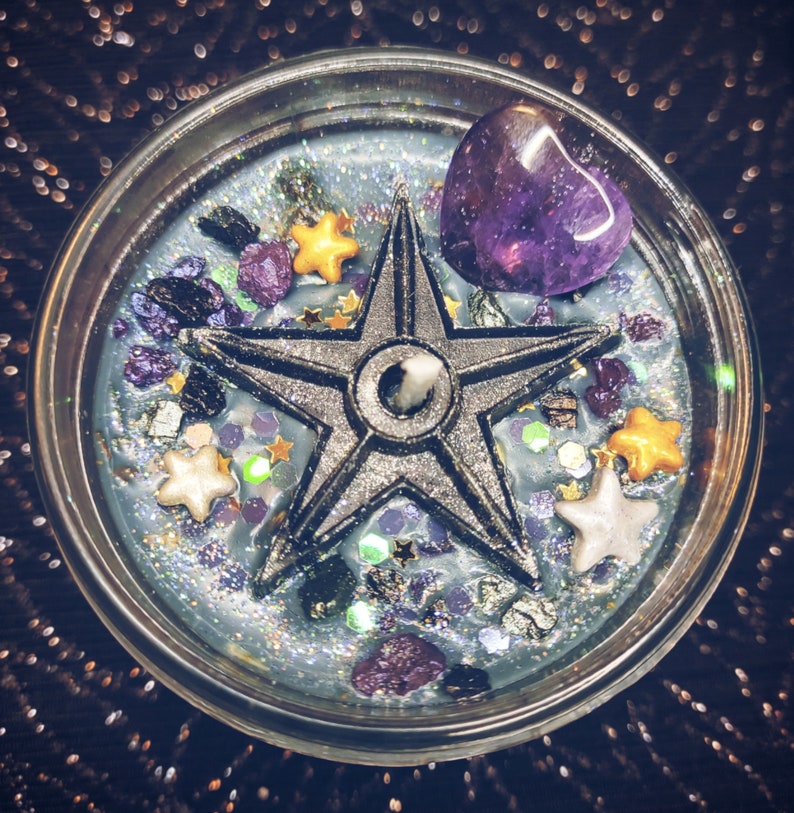 May include: A teal blue candle with a five-pointed star, purple amethyst crystal, and gold glitter. The candle is in a clear glass jar.