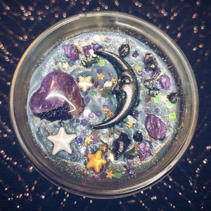 May include: A clear glass jar candle with a blue wax base and a variety of decorative elements, including a crescent moon, a purple heart, and glitter.