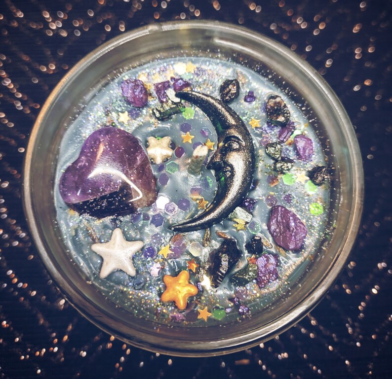 May include: A clear glass jar candle with a blue wax base and a variety of decorative elements, including a crescent moon, a purple heart, and glitter.