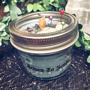 May include: A blue-green candle in a mason jar with a silver lid. The candle is decorated with small stones, a wooden stick, and colorful pins. The text "Return To Senda" is printed on the jar.