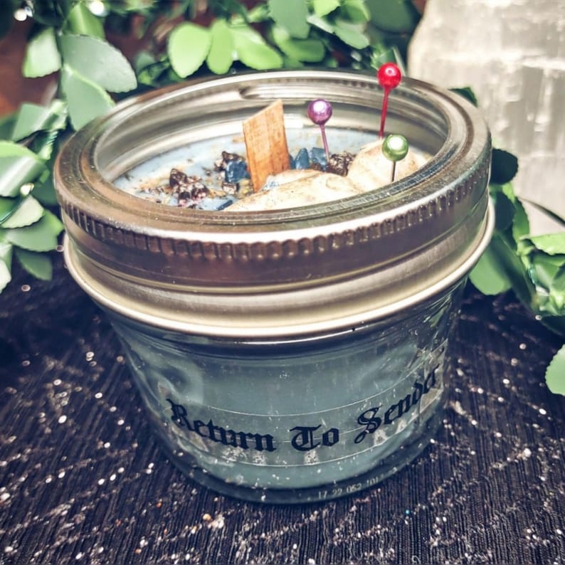May include: A blue-green candle in a mason jar with a silver lid. The candle is decorated with small stones, a wooden stick, and colorful pins. The text "Return To Senda" is printed on the jar.