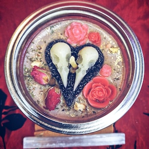 Può includere: Una candela in vetro a forma di cuore nero con due accenti di teschio bianco. La candela è circondata da rose rosse e rosa e altre piante.