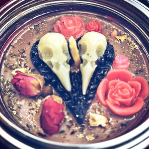 Puede incluir: Una vela negra en forma de coraz&oacute;n con dos calaveras blancas y rosas rojas. La vela est&aacute; en un frasco de vidrio con una tapa plateada.