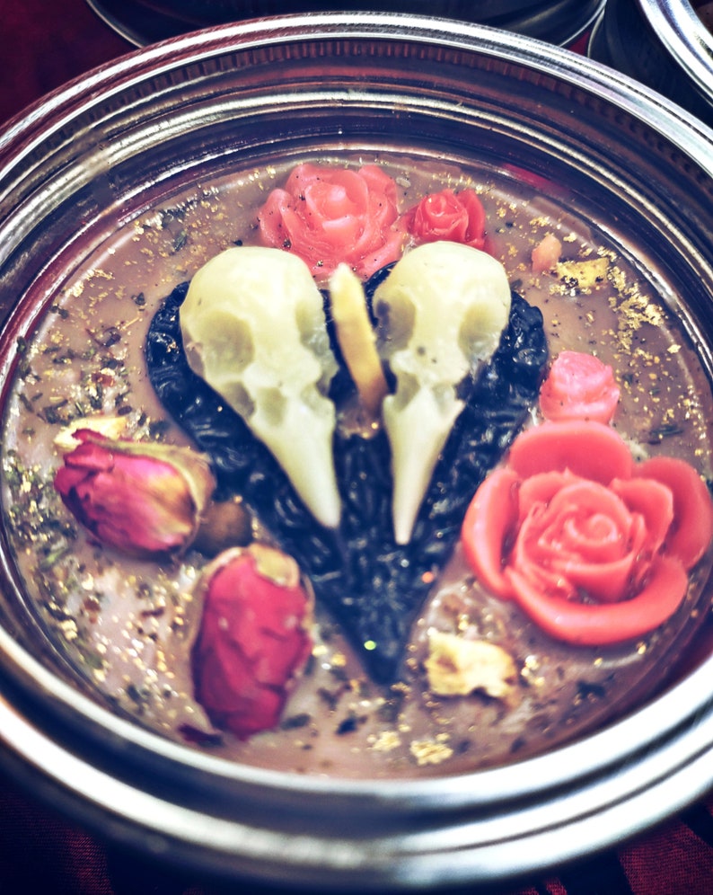 Puede incluir: Una vela negra en forma de coraz&oacute;n con dos calaveras blancas y rosas rojas. La vela est&aacute; en un frasco de vidrio con una tapa plateada.