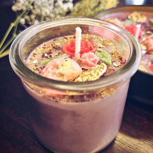 May include: A purple candle with a glass jar and a white wick. The candle is decorated with gold glitter, pink and red flowers, and green leaves.