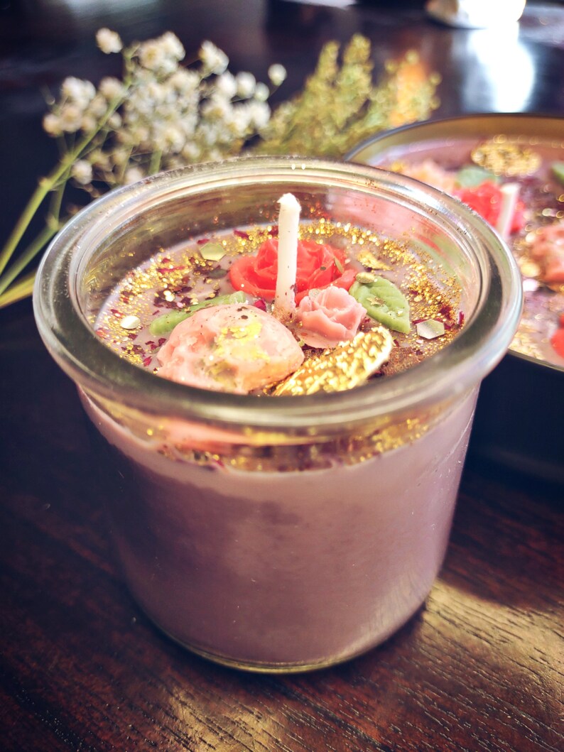 May include: A purple candle with a glass jar and a white wick. The candle is decorated with gold glitter, pink and red flowers, and green leaves.
