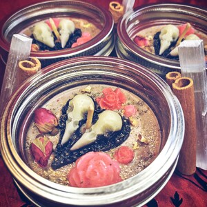 Puede incluir: Tres frascos de vidrio con tapas, llenos de una vela de cera marr&oacute;n oscuro. La vela tiene una forma de coraz&oacute;n negro con dos calaveras blancas en su interior. El coraz&oacute;n est&aacute; rodeado de rosas rosas y purpurina dorada.