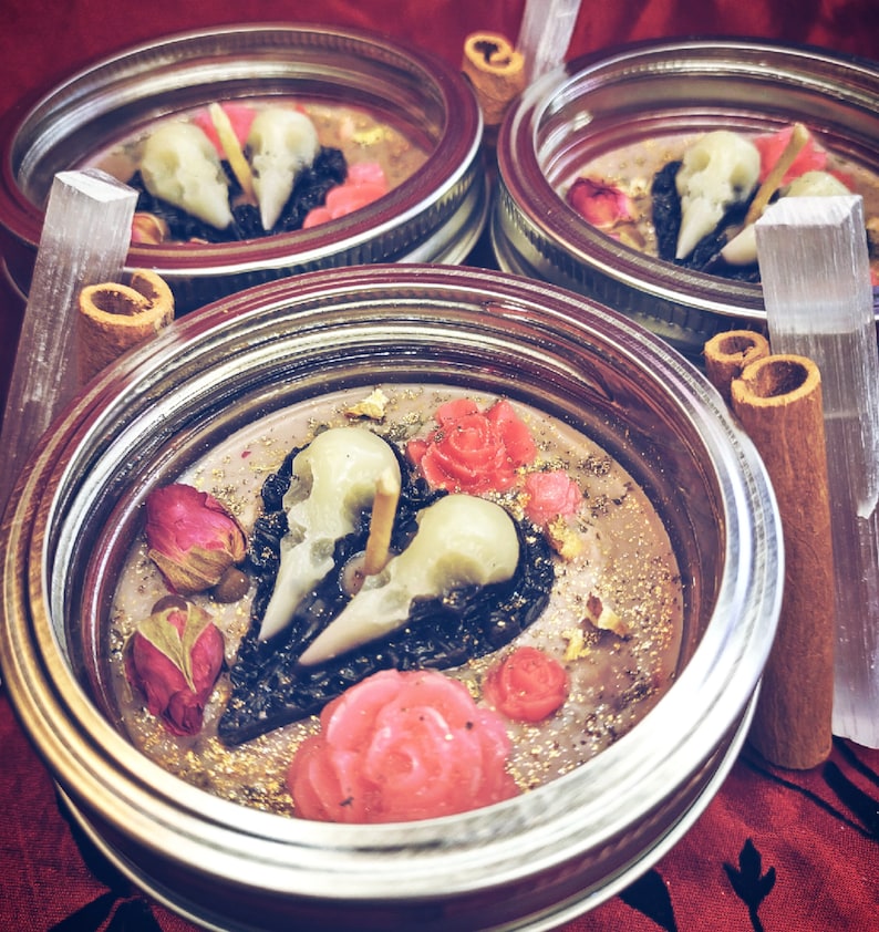 Puede incluir: Tres frascos de vidrio con tapas, llenos de una vela de cera marr&oacute;n oscuro. La vela tiene una forma de coraz&oacute;n negro con dos calaveras blancas en su interior. El coraz&oacute;n est&aacute; rodeado de rosas rosas y purpurina dorada.