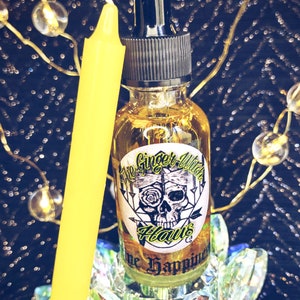 May include: A clear glass bottle with a black dropper top and a yellow label that reads "The Ginger Witch Haus" and "Hope Happiness". The bottle is sitting on a decorative glass base with a yellow candle next to it.