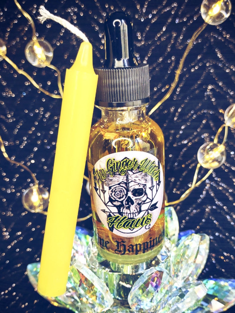 May include: A clear glass bottle with a black dropper top and a yellow label that reads "The Ginger Witch Haus" and "Hope Happiness". The bottle is sitting on a decorative glass base with a yellow candle next to it.