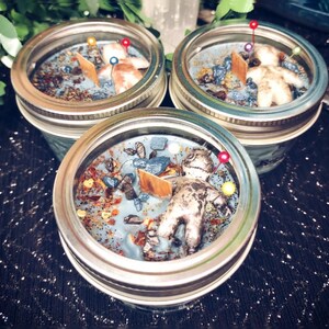 May include: Three mason jar candles with a light blue wax, decorated with small stones, wooden pieces, and a voodoo doll. Each candle has a metal lid and is set on a dark, sparkly surface.