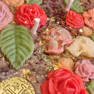 May include: A purple candle with a floral and skull design. The candle features pink, red, and green roses, leaves, and skulls. The candle is decorated with gold glitter.