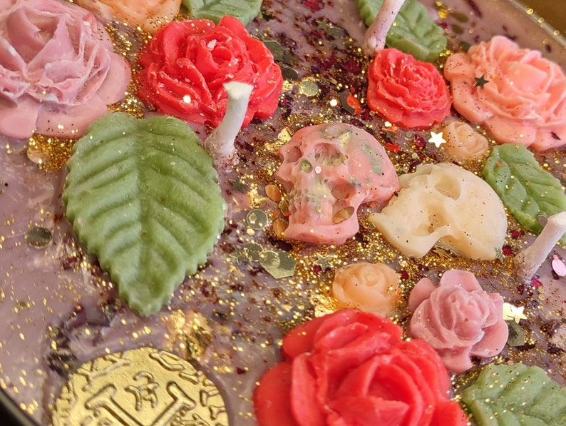 May include: A purple candle with a floral and skull design. The candle features pink, red, and green roses, leaves, and skulls. The candle is decorated with gold glitter.