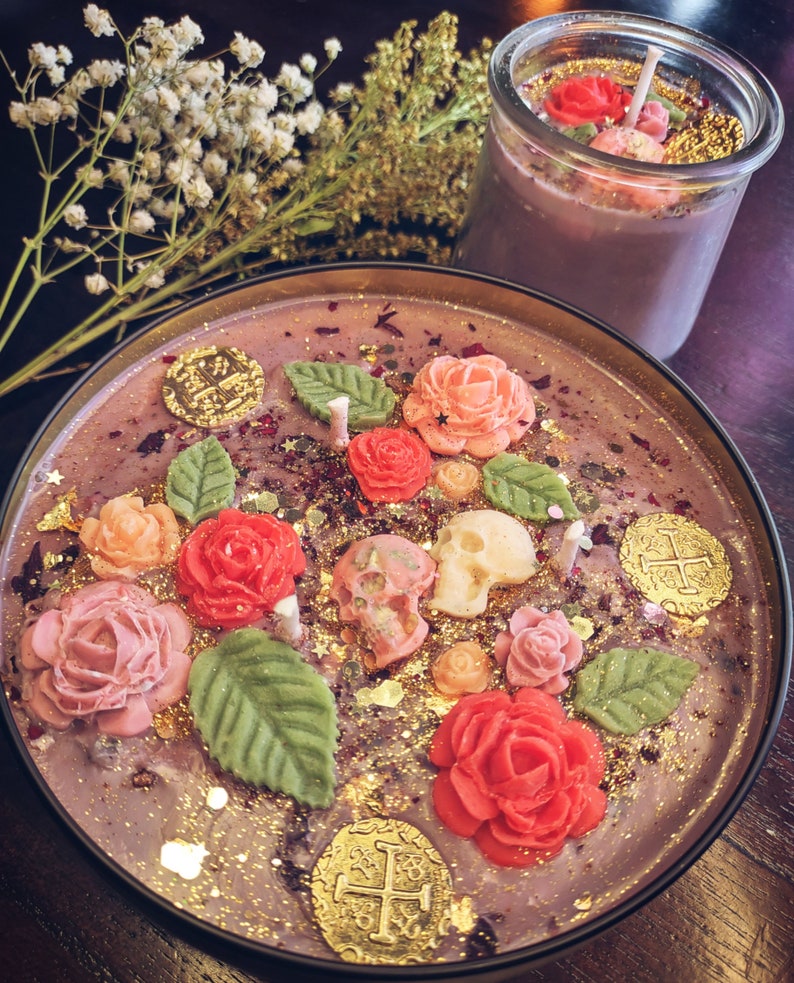 May include: Purple candle with gold glitter, gold coins, and pink and red roses. The candle is in a glass jar with a black rim.