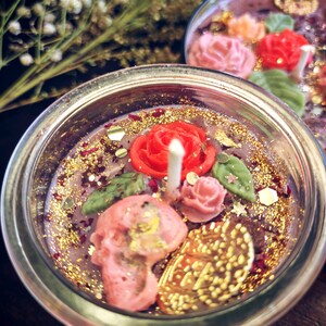 May include: A glass jar candle with a pink and red floral design, gold glitter, and a skull. The candle is surrounded by gold glitter and has a pink and red floral design.