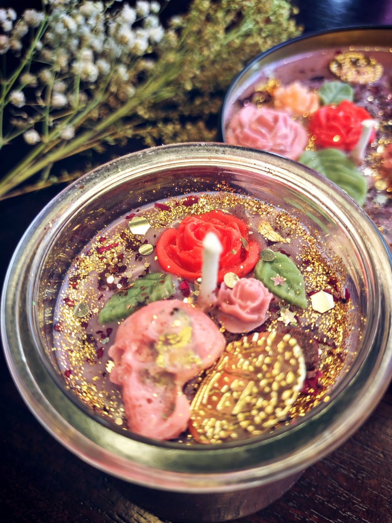 May include: A glass jar candle with a pink and red floral design, gold glitter, and a skull. The candle is surrounded by gold glitter and has a pink and red floral design.