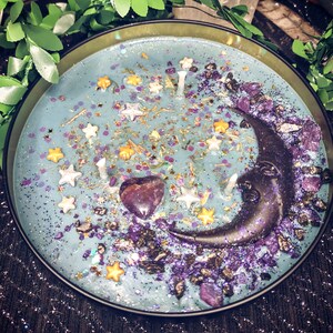 May include: A blue candle with a crescent moon design, purple and gold glitter, and a purple heart-shaped crystal. The candle is in a black metal container.