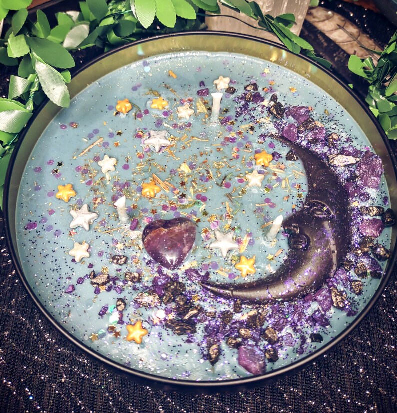 May include: A blue candle with a crescent moon design, purple and gold glitter, and a purple heart-shaped crystal. The candle is in a black metal container.
