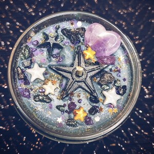 May include: A blue candle with a silver five-pointed star, purple heart, and gold and white stars embedded in the wax. The candle is surrounded by glitter and small black and green stones.