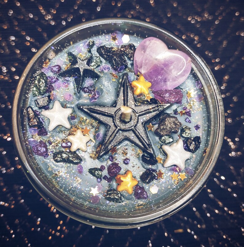 May include: A blue candle with a silver five-pointed star, purple heart, and gold and white stars embedded in the wax. The candle is surrounded by glitter and small black and green stones.