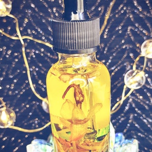 May include: A clear glass bottle with a black dropper top filled with a yellow liquid and a dried flower inside. The bottle is sitting on a clear glass base.