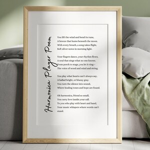 May include: A framed print featuring a poem titled "Harmonica Player Poem" in elegant script. The poem is printed in black on a white background, encased in a light wood frame. The artwork is displayed on a gray sofa with pillows and a green blanket.