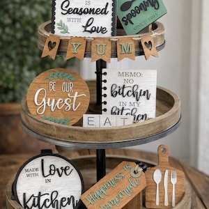May include: A three-tiered wooden tray with decorative kitchen-themed signs. The signs feature phrases like "This Kitchen is Seasoned with Love," "Be Our Guest," and "Happiness is Homemade." The tray includes a spoon-shaped sign and a cutting board with utensils.