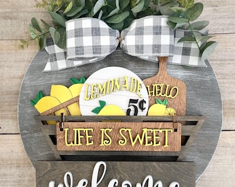 Puede incluir: Un letrero redondo de madera con fondo gris y un letrero de bienvenida blanco. El letrero está decorado con un lazo a cuadros blanco y negro, hojas verdes y un letrero de madera que dice "Lemonade 5¢ Hello". También hay un letrero de madera que dice "Life is Sweet" y una cesta con limones.