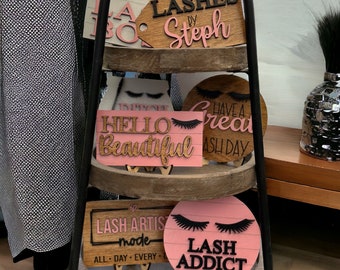 May include: A tiered wooden stand with five pink and brown wooden signs with eyelash designs and text. The signs read "LASHES by Steph", "HELLO Beautiful", "HAVE A GREAT LASH DAY", "LASH ARTIST mode ALL DAY EVERY", and "LASH ADDICT".
