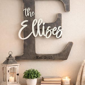 May include: A large wooden letter "E" with the name "the Ellises" in white script. The year "Est. 2025" is below. A decorative lantern, potted plant, books, and candle sit on a wooden table.