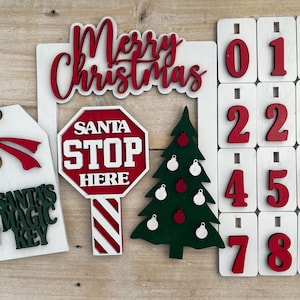 May include: A white wooden Christmas countdown calendar with red numbers, a red and white striped stop sign with the words "Santa Stop Here", a green Christmas tree with red ornaments, a white tag with a gold key and the words "Santa's Magic Key", and a white sign with the words "Merry Christmas".
