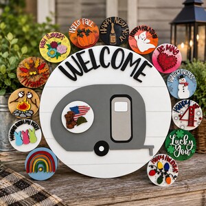 May include: A round white wooden welcome sign featuring a gray camper trailer in the center. The sign includes twelve interchangeable circular wooden icons representing various holidays and seasons, such as a pumpkin, ghost, snowman, and rainbow.