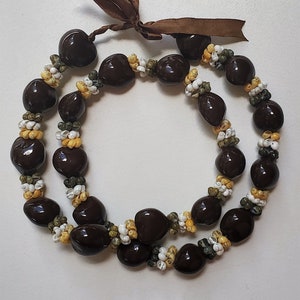 May include: A brown seed lei with white and yellow shell accents. The lei is made of two strands of seeds and is tied together with a brown ribbon.