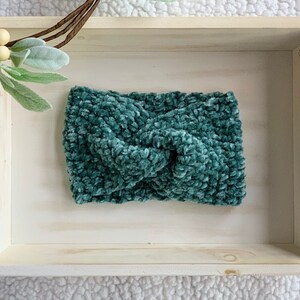 Puede incluir: Una diadema de crochet azul turquesa con un diseño de nudo retorcido. La diadema se muestra en una bandeja de madera.