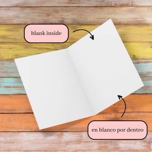 May include: An open, blank white card with the text "blank inside" and "en blanco por dentro" on a multicolored wooden surface. The card is angled, revealing the inside pages. The background features a mix of orange, yellow, and blue wood.