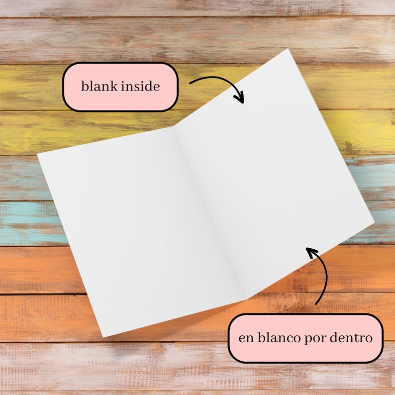 May include: An open, blank white card with the text "blank inside" and "en blanco por dentro" on a multicolored wooden surface. The card is angled, revealing the inside pages. The background features a mix of orange, yellow, and blue wood.
